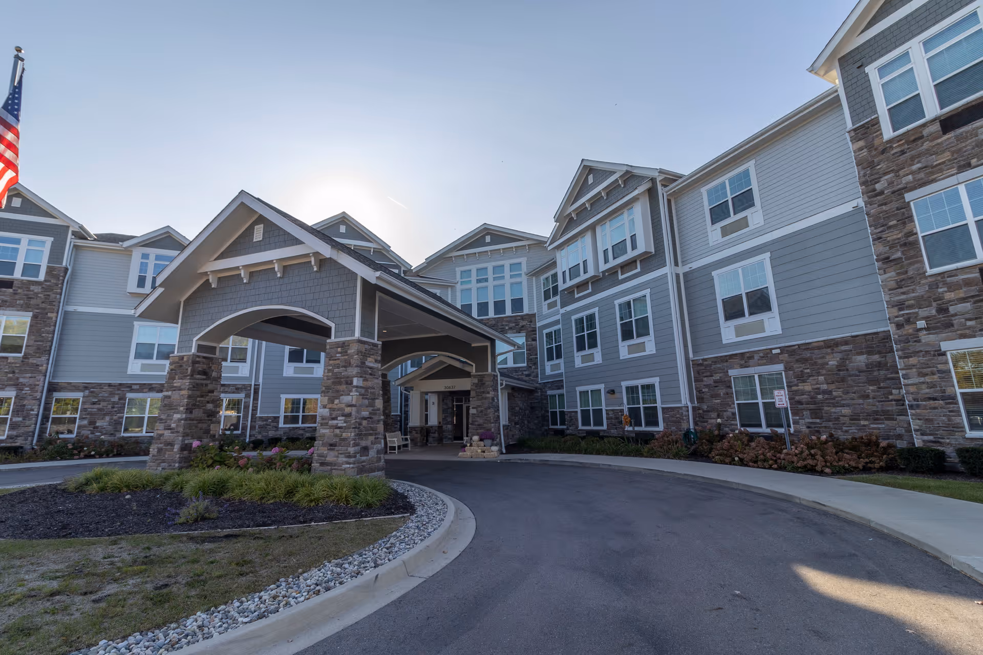 Exterior view of a multi-story senior living facility building with a covered entrance supported by stone pillars, landscaped greenery, and an American flag on the left side.