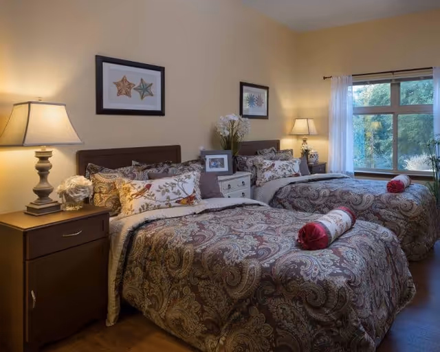 A cozy bedroom with two twin beds featuring patterned bedspreads and decorative pillows. Each bed has a cylindrical bolster pillow in red and white. Between the beds is a small white nightstand with a vase of flowers and a framed photo. On either side of the beds are wooden nightstands with lamps. The room has beige walls, two framed starfish pictures above the beds, and a large window with sheer white curtains letting in natural light and showing greenery outside.