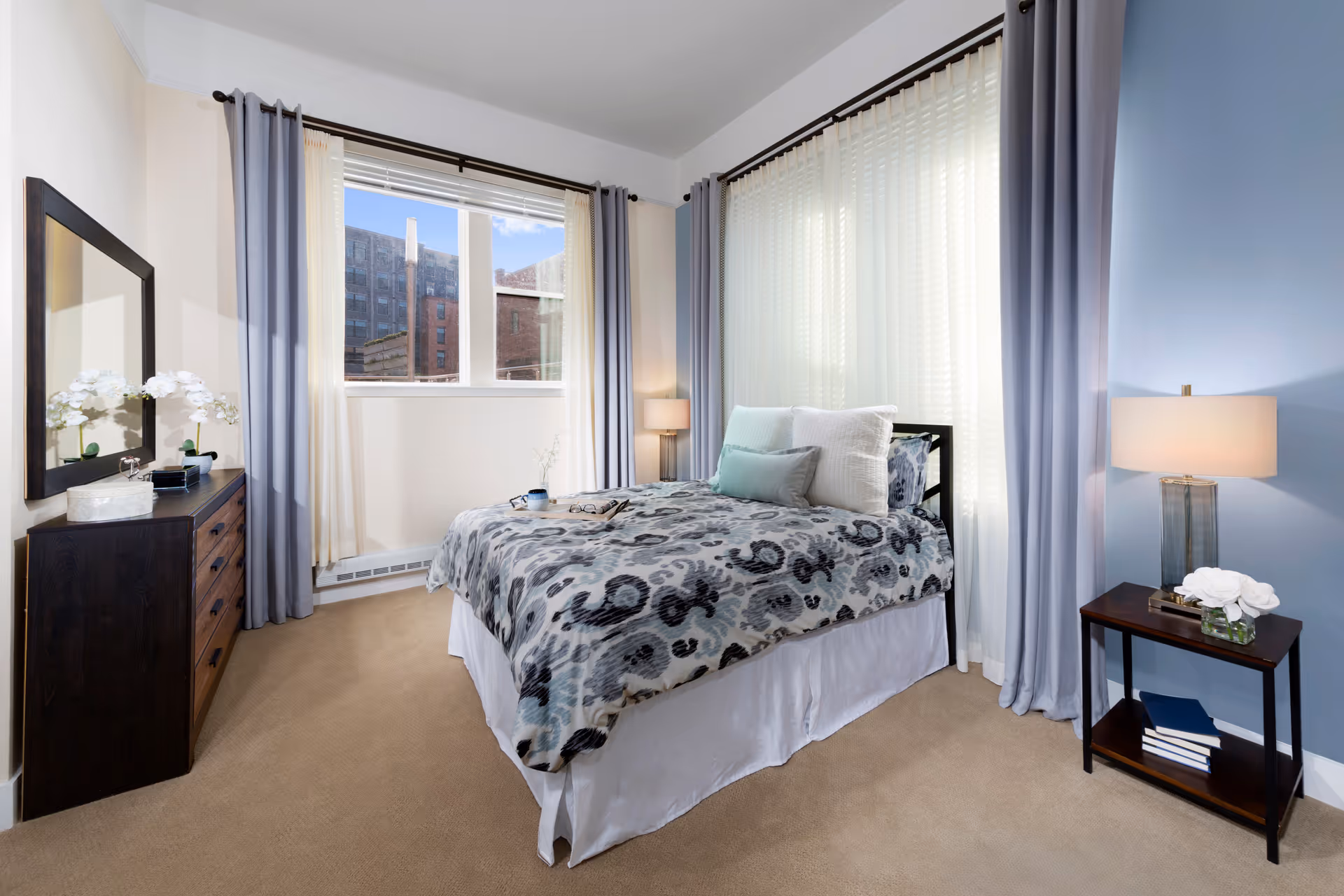 A bright, neatly staged bedroom featuring a made bed with a patterned duvet, a dresser with mirror, nightstands with lamps, and large windows with curtains.