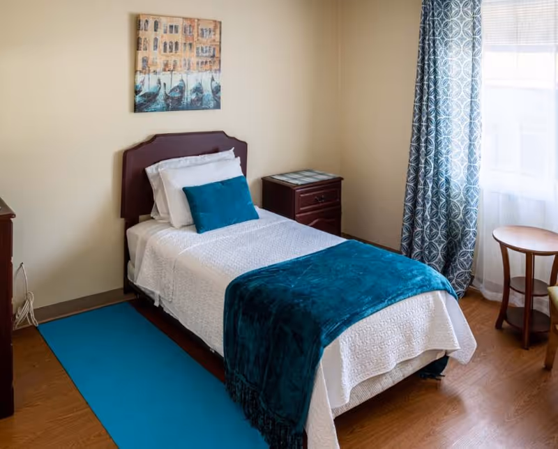 A tidy single bedroom with a bed dressed in white linens and teal throw and pillow, a nightstand, patterned curtains, and wall art.