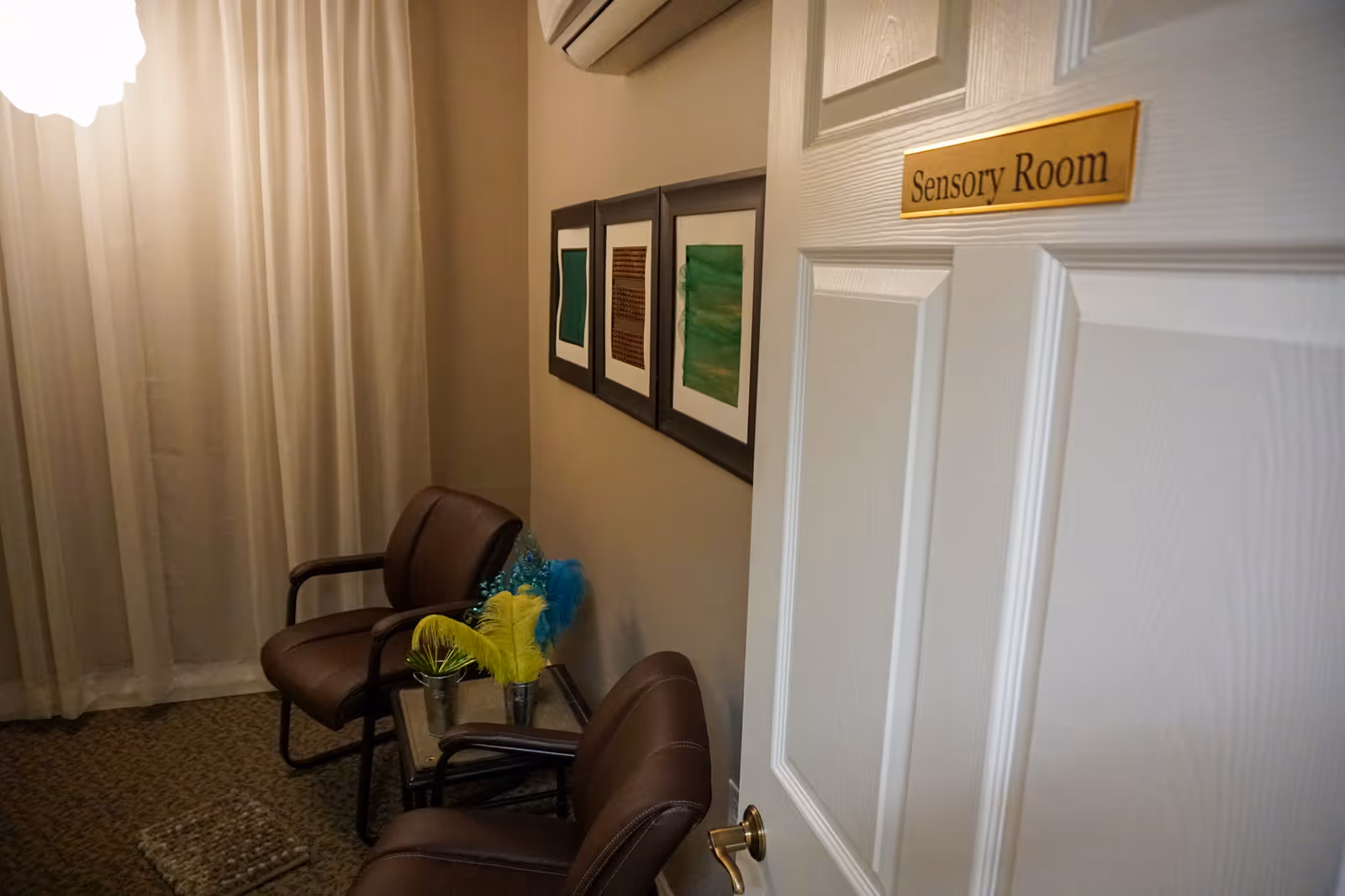 Interior view of a sensory room with two brown chairs, a small table holding colorful feather decorations, three framed abstract artworks on the wall, beige curtains, and a white door with a gold sign reading 'Sensory Room'.