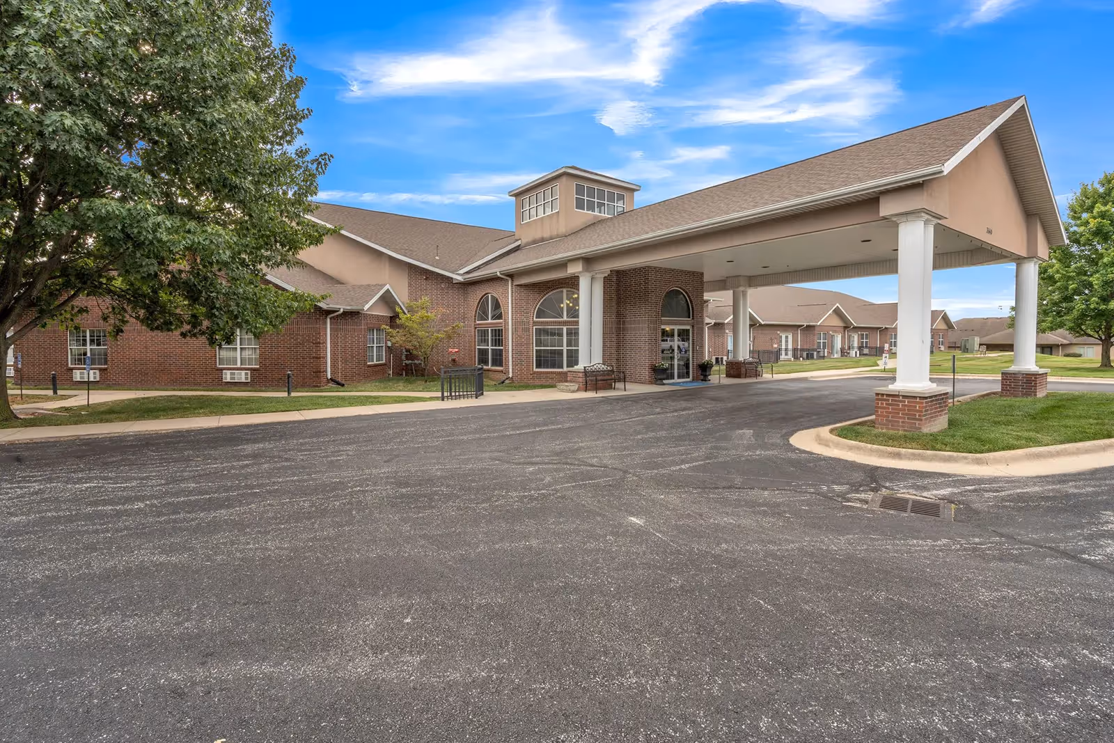 Brick single-story senior living building with a covered entrance canopy, paved driveway, and surrounding lawn under a blue sky.