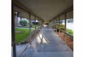 Covered outdoor walkway with a concrete path and supporting columns flanked by landscaped beds leading toward a building.