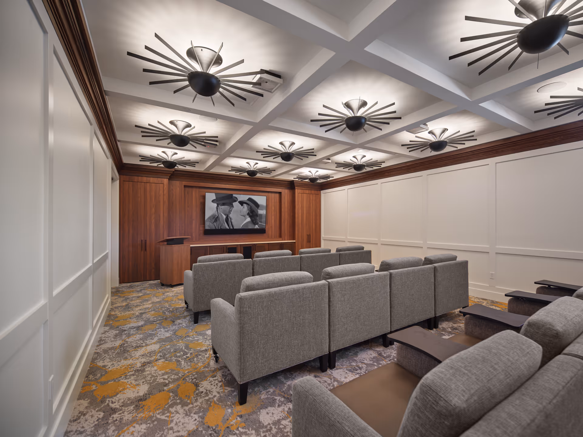 A cozy movie screening room with rows of gray upholstered chairs facing a large screen displaying a black and white film. The room features a patterned carpet with yellow and gray tones, white paneled walls, a coffered ceiling with multiple modern black and silver ceiling fans, and wooden cabinetry at the front.