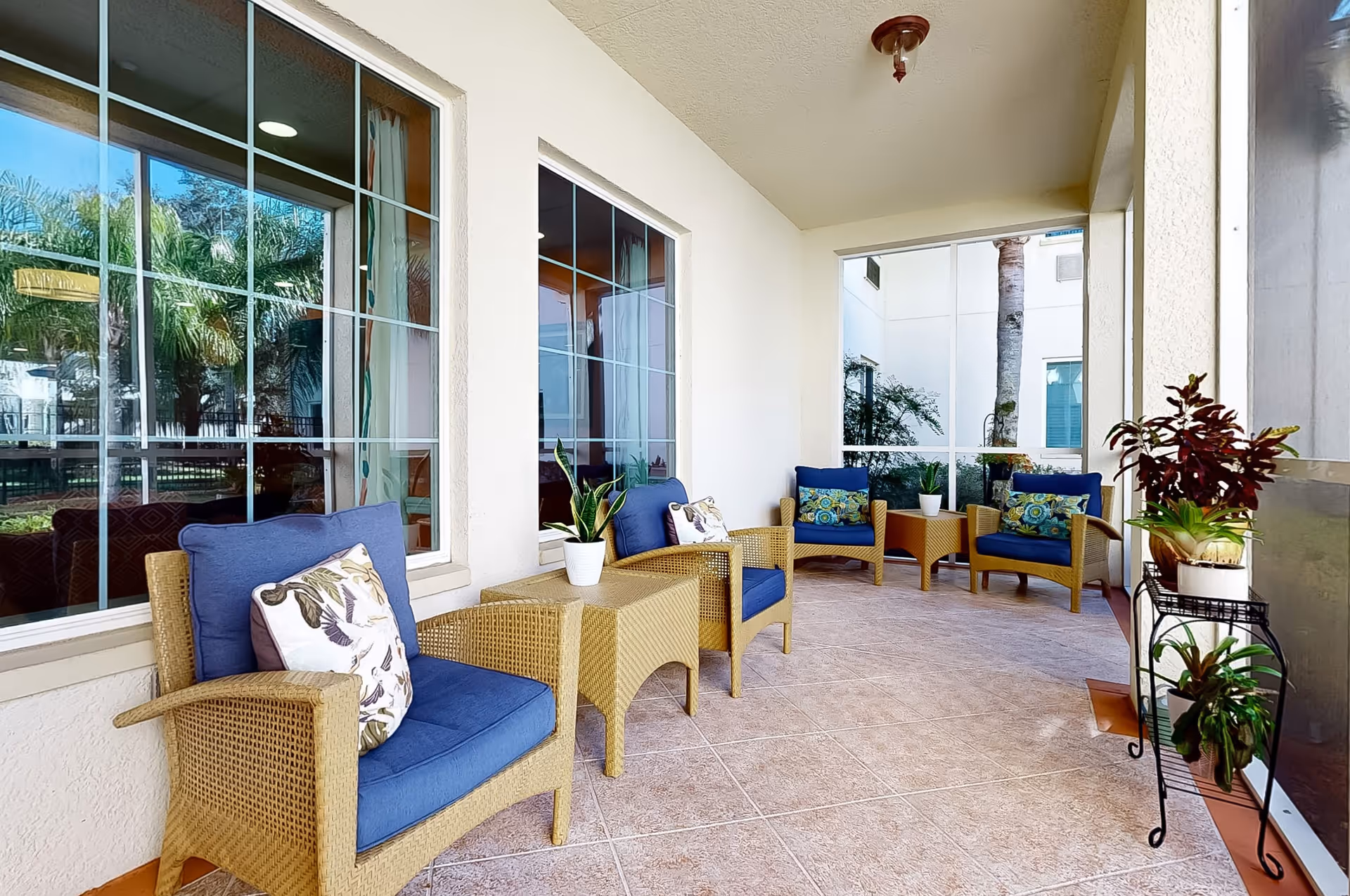 Covered outdoor patio area with wicker chairs featuring blue cushions and decorative pillows, small tables with potted plants, and large windows reflecting palm trees and greenery outside.