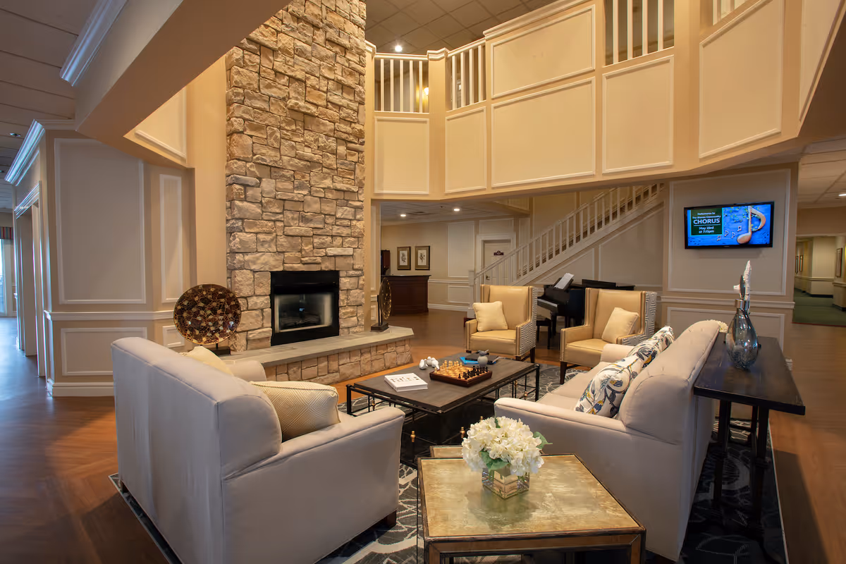 A cozy assisted living facility common area with beige sofas and armchairs arranged around a coffee table with a chessboard. There is a stone fireplace in the background, a piano near a staircase, and a wall-mounted TV displaying an event. The room has warm lighting and elegant wall paneling.
