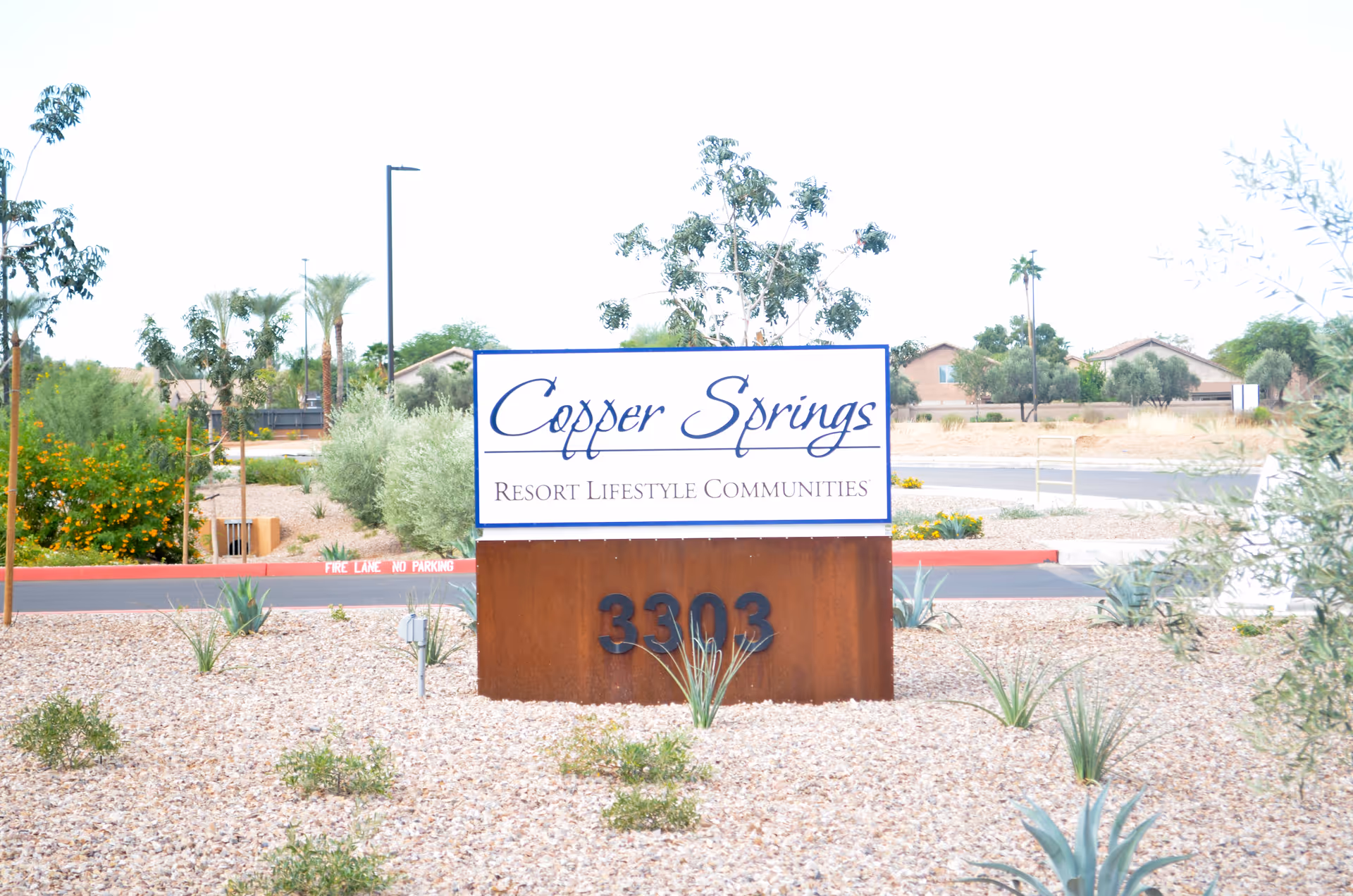 Outdoor view of a sign for Copper Springs Resort Lifestyle Communities with the address number 3303 displayed below. The sign is surrounded by desert landscaping with small plants and gravel, and there are trees and houses visible in the background.