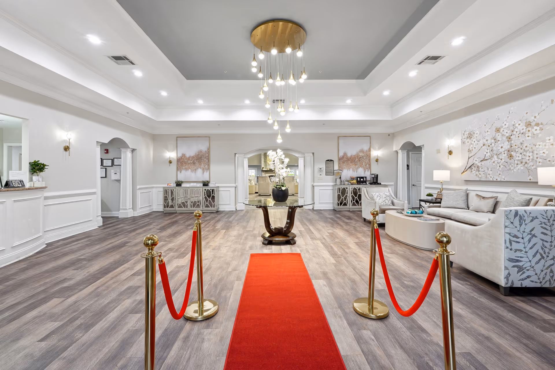 Spacious and elegant lobby area with a red carpet flanked by gold stanchions and red ropes, a round glass table with a flower arrangement in the center, modern chandelier hanging from a recessed ceiling, light wood flooring, white walls with decorative molding, two matching sideboards with artwork above them, and a seating area with a sofa and armchairs on the right side.