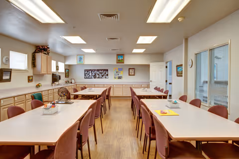 A well-lit room with multiple rectangular tables arranged in rows, surrounded by maroon chairs. The room has light-colored walls with several framed pictures and bulletin boards. There are cabinets along the back and left walls, and fluorescent ceiling lights illuminate the space. A door is visible at the back right, and windows are on the right wall.