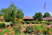 A well-maintained garden with colorful flowers and green shrubs in front of a single-story building with a brown roof. An American flag is flying on a flagpole to the right under a clear blue sky.
