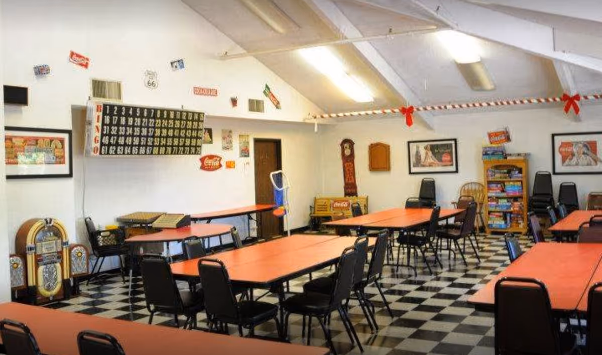A senior community activity room with red-topped tables and black chairs, a large bingo board on the wall, game shelves and a jukebox on a black-and-white checkered floor.