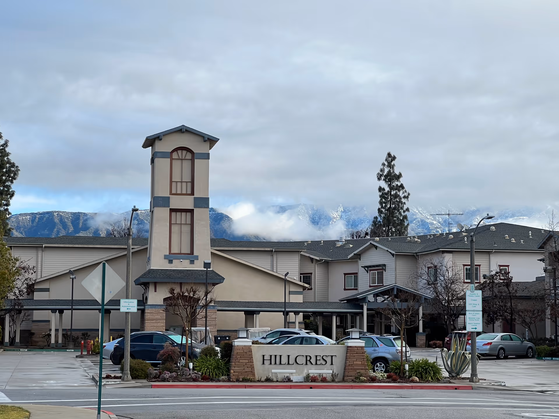 Exterior view of the Hillcrest senior living facility with a multi-story building, a tower-like structure in the center, several parked cars, and mountains in the background under a cloudy sky.
