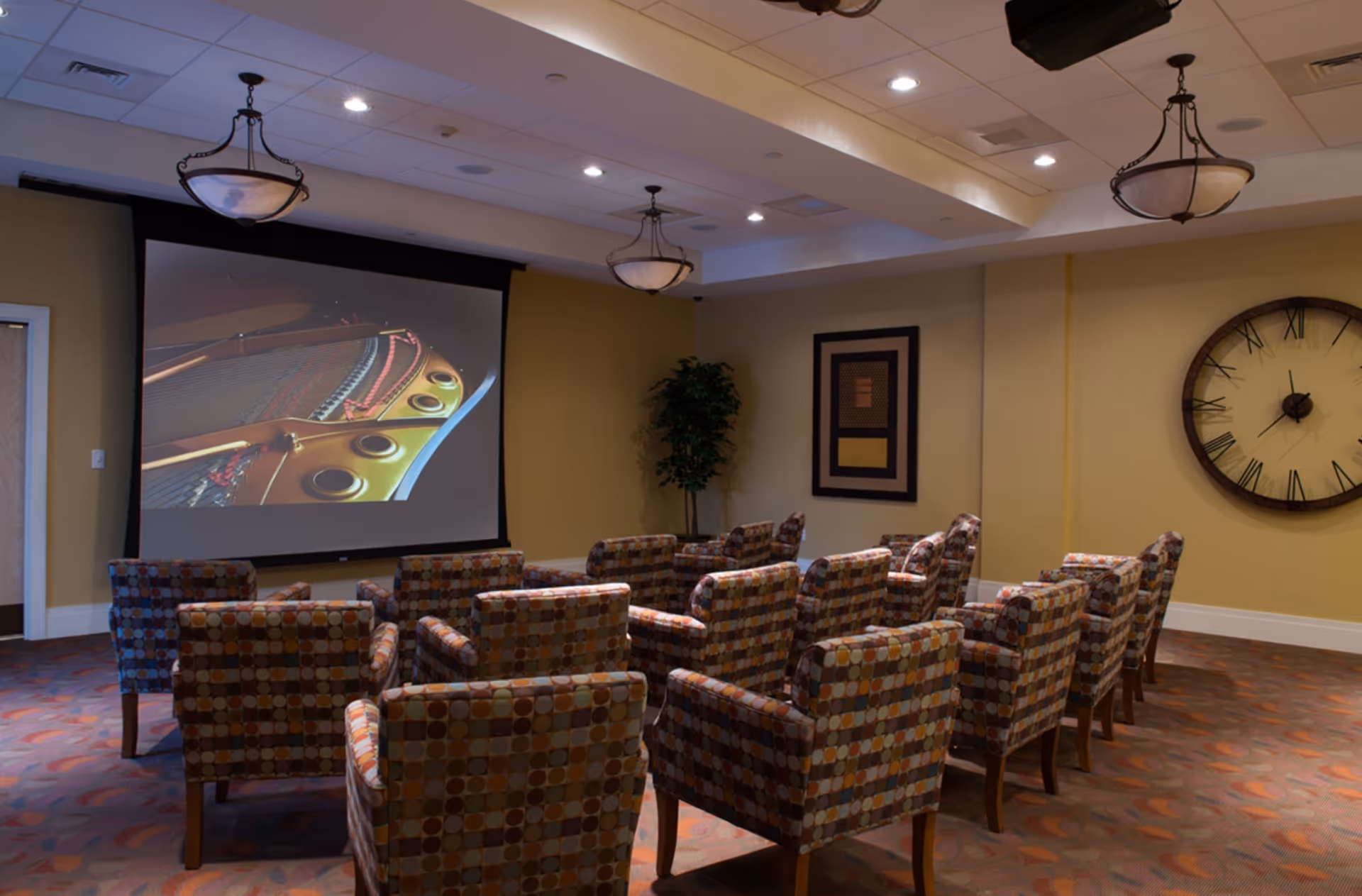 A small theater or media room with multiple patterned armchairs arranged in rows facing a large projection screen displaying a close-up image of a piano's interior. The room has warm yellow walls, a large round clock with Roman numerals on the right wall, a framed artwork, a potted plant in the corner, and ceiling lights with decorative fixtures.