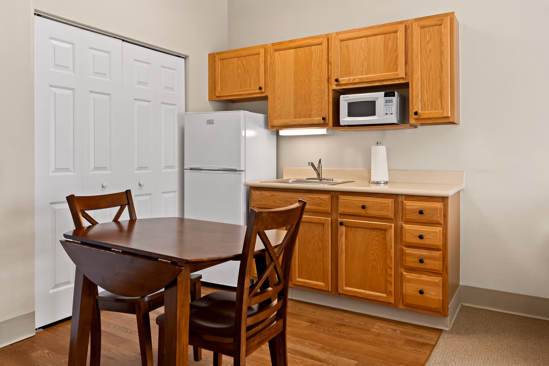 A small kitchen area with wooden cabinets, a white refrigerator, a microwave, a sink, and a paper towel holder. In front of the kitchen is a wooden dining table with two matching chairs. The floor is wood and there is a white double-door closet in the background.