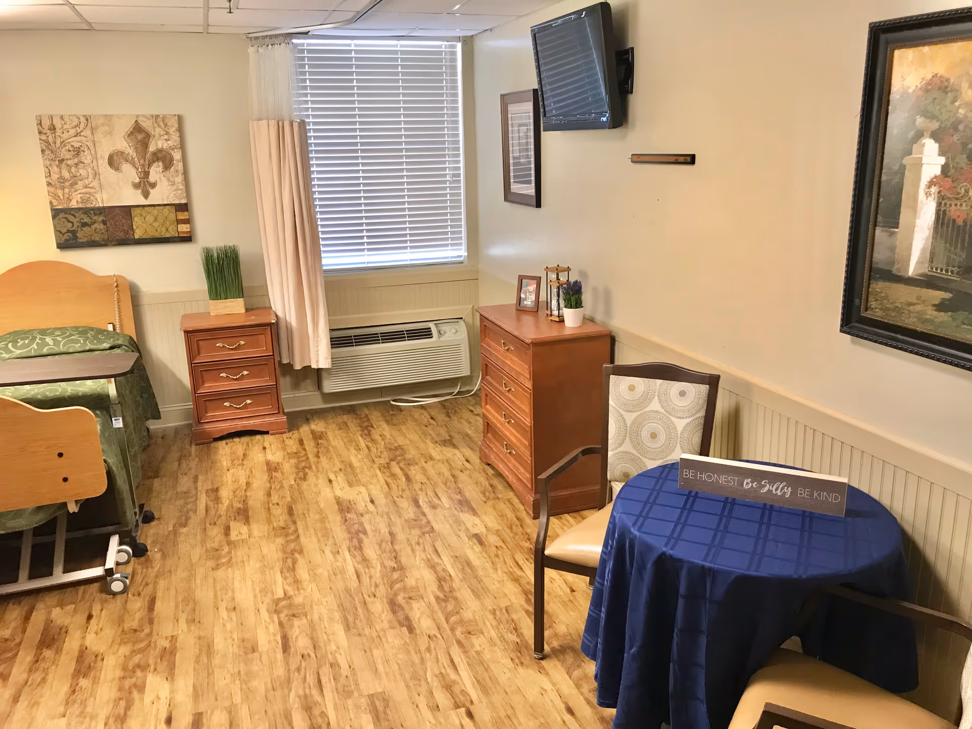 Small furnished resident bedroom with a bed, two dressers, a wall-mounted TV, and a round table with chairs.