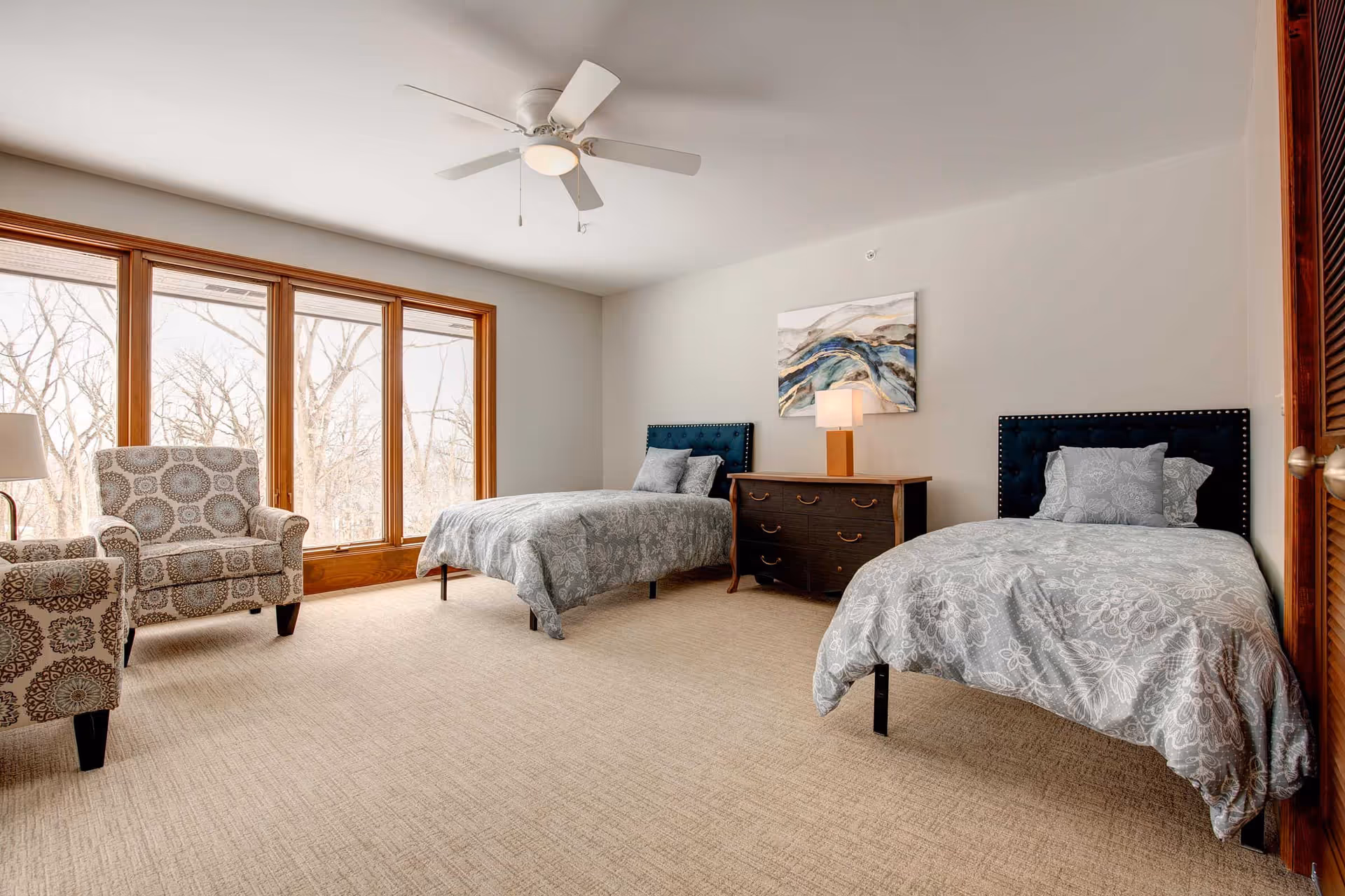 Spacious bedroom with two twin beds, a dresser and patterned armchairs beside large windows.