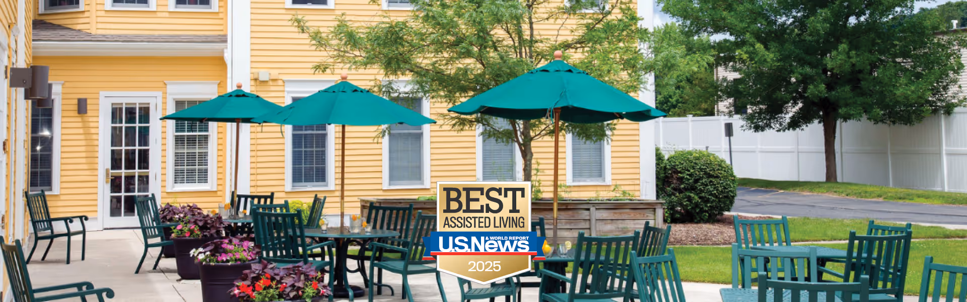 Outdoor patio with green tables, chairs, and umbrellas in front of a yellow senior living building with a 'BEST ASSISTED LIVING U.S. NEWS 2025' badge.