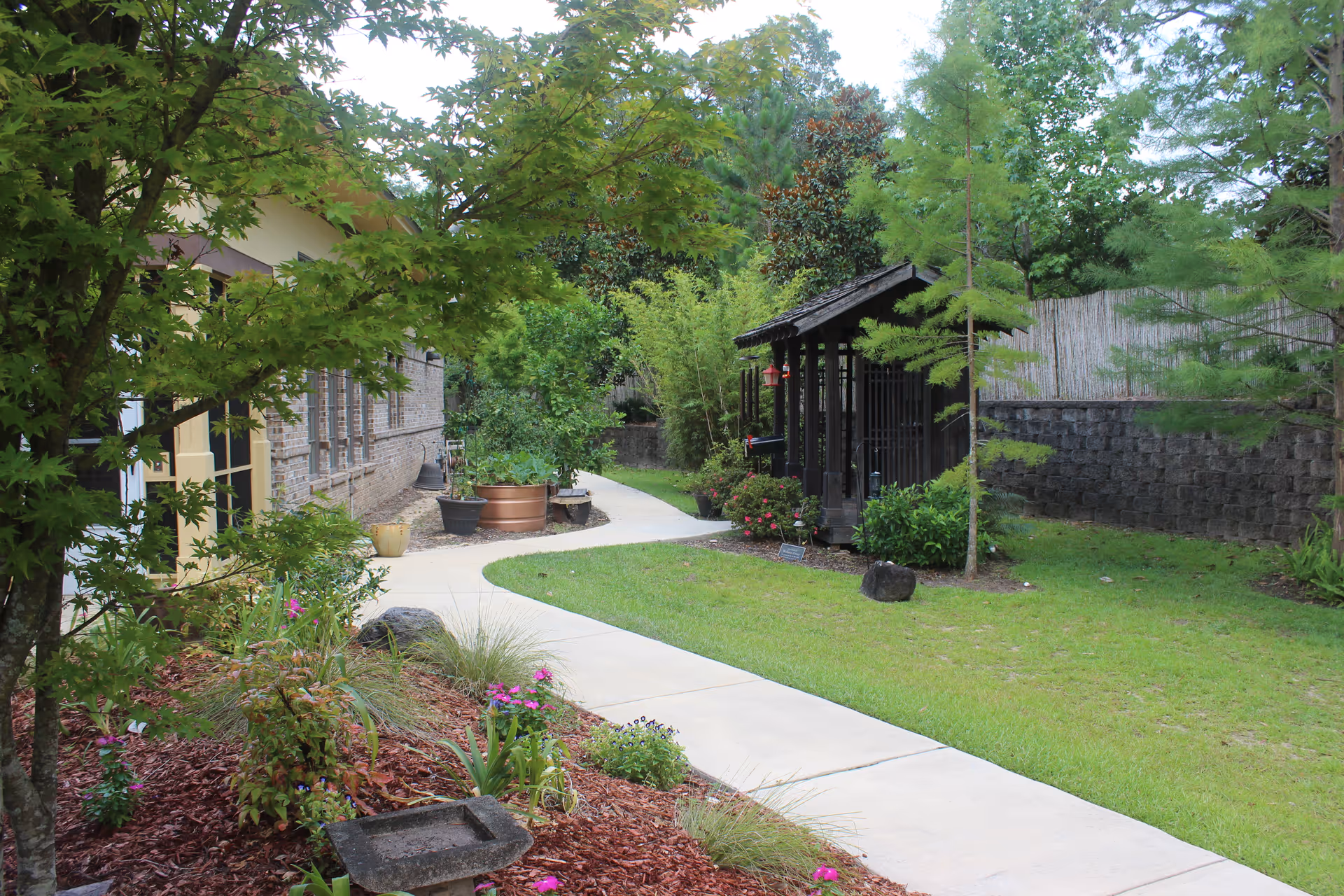 A landscaped outdoor garden area with a curved concrete pathway, green grass, various plants and trees, a small wooden gazebo, and a brick building on the left side.