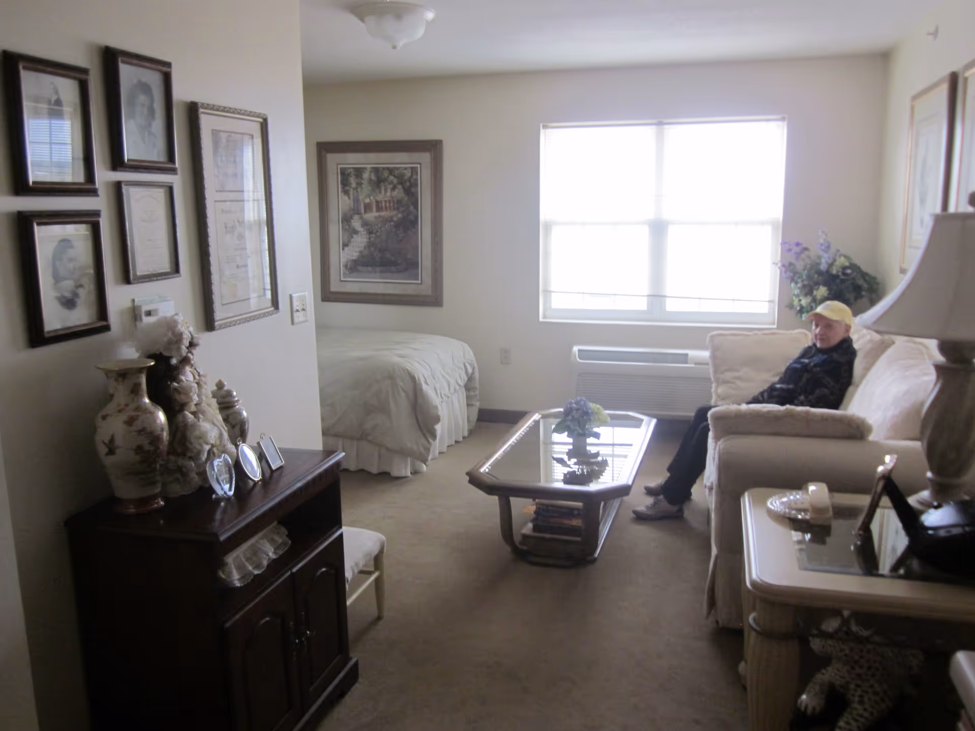 Studio apartment living area with a bed in the background, a sofa with a person seated by a window, a coffee table, and a cabinet with framed photos and decorative vases.