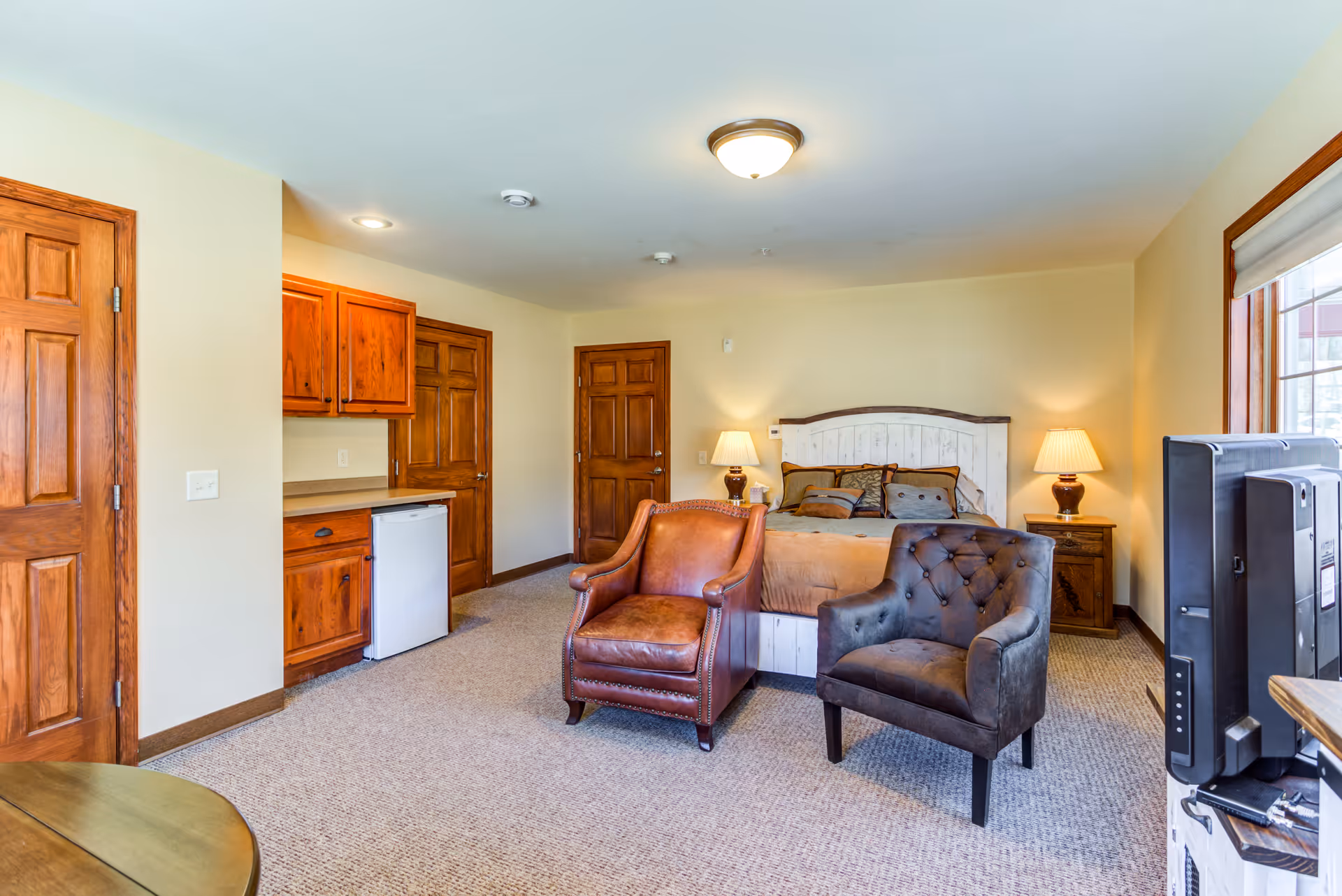 A cozy bedroom in an assisted living facility featuring a bed with pillows and a headboard, two armchairs in front of the bed, wooden nightstands with lamps on each side, a small kitchenette area with cabinets and a mini fridge, and a television on a stand near a window with blinds.
