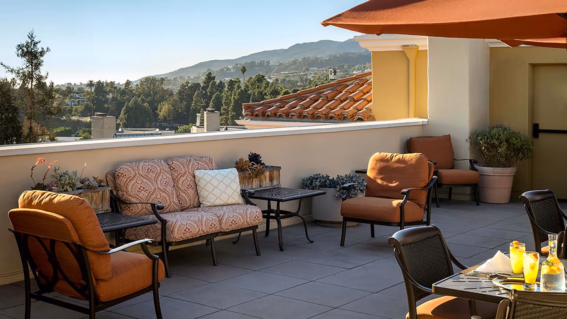 Outdoor patio area with cushioned chairs and a loveseat, small tables, potted plants, and a table set with drinks and napkins under a large umbrella, with a scenic view of trees and mountains in the background.