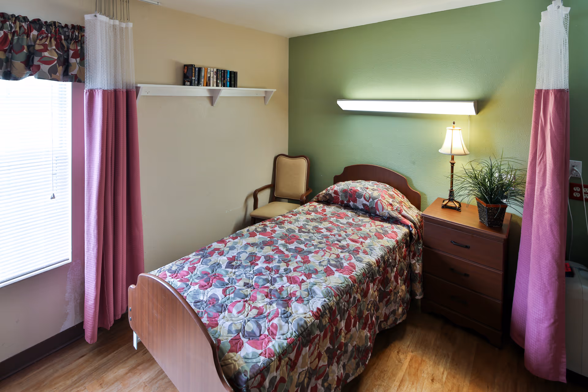 A small bedroom with a single bed covered in a floral quilt, a bedside dresser with a lamp and plant, a chair, and pink curtains by the window.