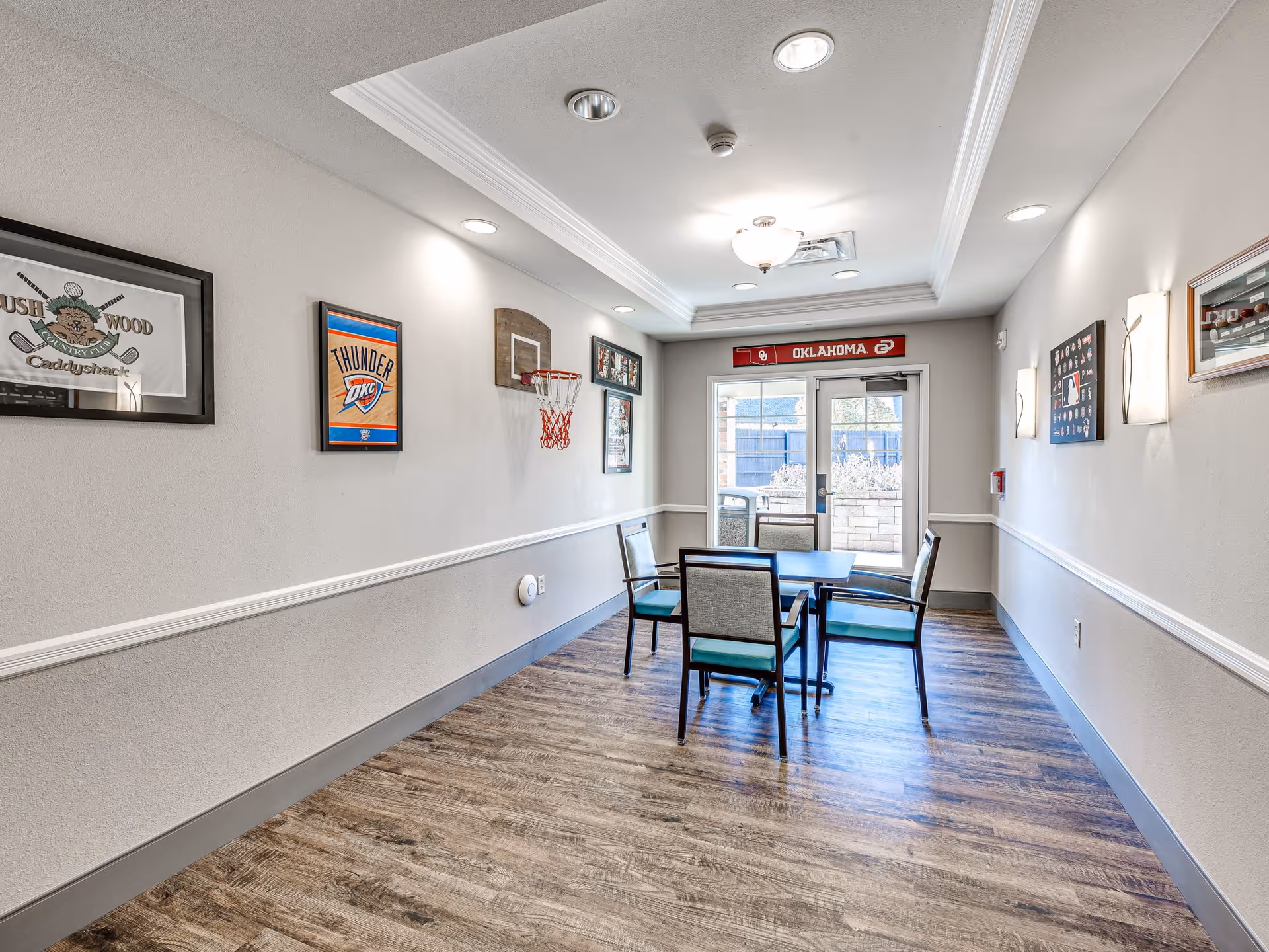 A small recreational room with a wooden floor and light gray walls. There is a square table with four chairs around it in the center of the room. On the left wall, there are framed sports memorabilia and a small basketball hoop. The back wall has a glass door leading outside, above which is a red Oklahoma sign. The room is well-lit with ceiling lights and wall sconces.