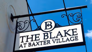 A decorative black metal hanging sign with scrollwork details displaying the text 'THE BLAKE AT BAXTER VILLAGE' against a clear blue sky background.