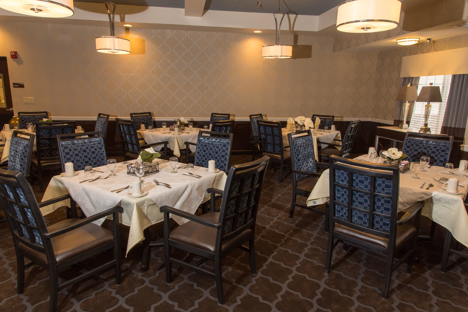 A dining room with multiple tables covered in white tablecloths, each set with cups, glasses, silverware, and small floral centerpieces. The room has patterned carpet, upholstered chairs with blue patterned backs, and soft lighting from ceiling fixtures and table lamps near a window.
