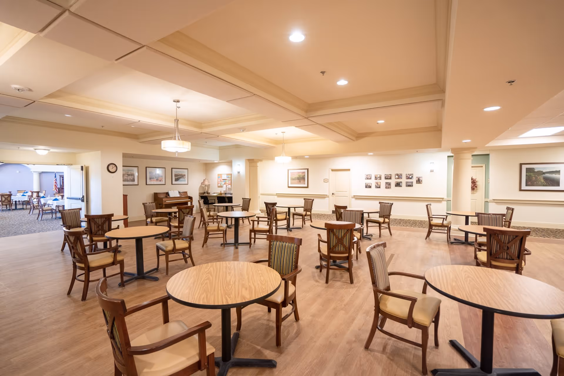 A spacious dining area with multiple round wooden tables and chairs arranged neatly on a wood floor. The room has a coffered ceiling with recessed lighting and hanging light fixtures. The walls are decorated with framed pictures and there is an open doorway leading to another room with more tables and chairs. The overall ambiance is warm and inviting.