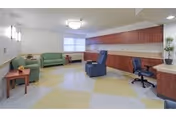 A spacious common area with a green sofa, matching loveseat, a blue recliner chair, and a black office chair. The room has a long wooden cabinet along one wall, a small wooden side table with a plant, and a large window with blinds. The floor has a patterned design with yellow and gray tiles, and the walls are painted white with modern light fixtures.