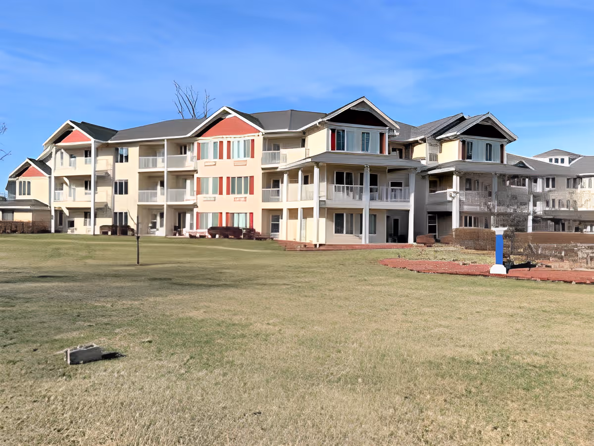 Exterior view of a multi-story senior living facility building with balconies and a large grassy lawn in front under a clear blue sky.