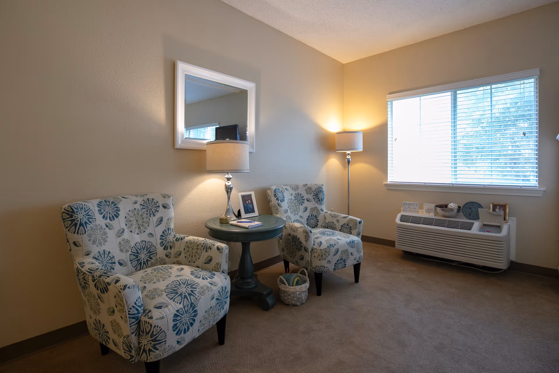 A cozy sitting area in a room with two patterned armchairs featuring blue and green floral designs, a round green side table between them with a lamp, a photo frame, and a notebook. A basket with yarn is on the floor next to one chair. A floor lamp stands near a window with white blinds, and a white air conditioning unit below the window holds decorative items including a clock and small framed pictures. A large mirror hangs on the wall above the side table.