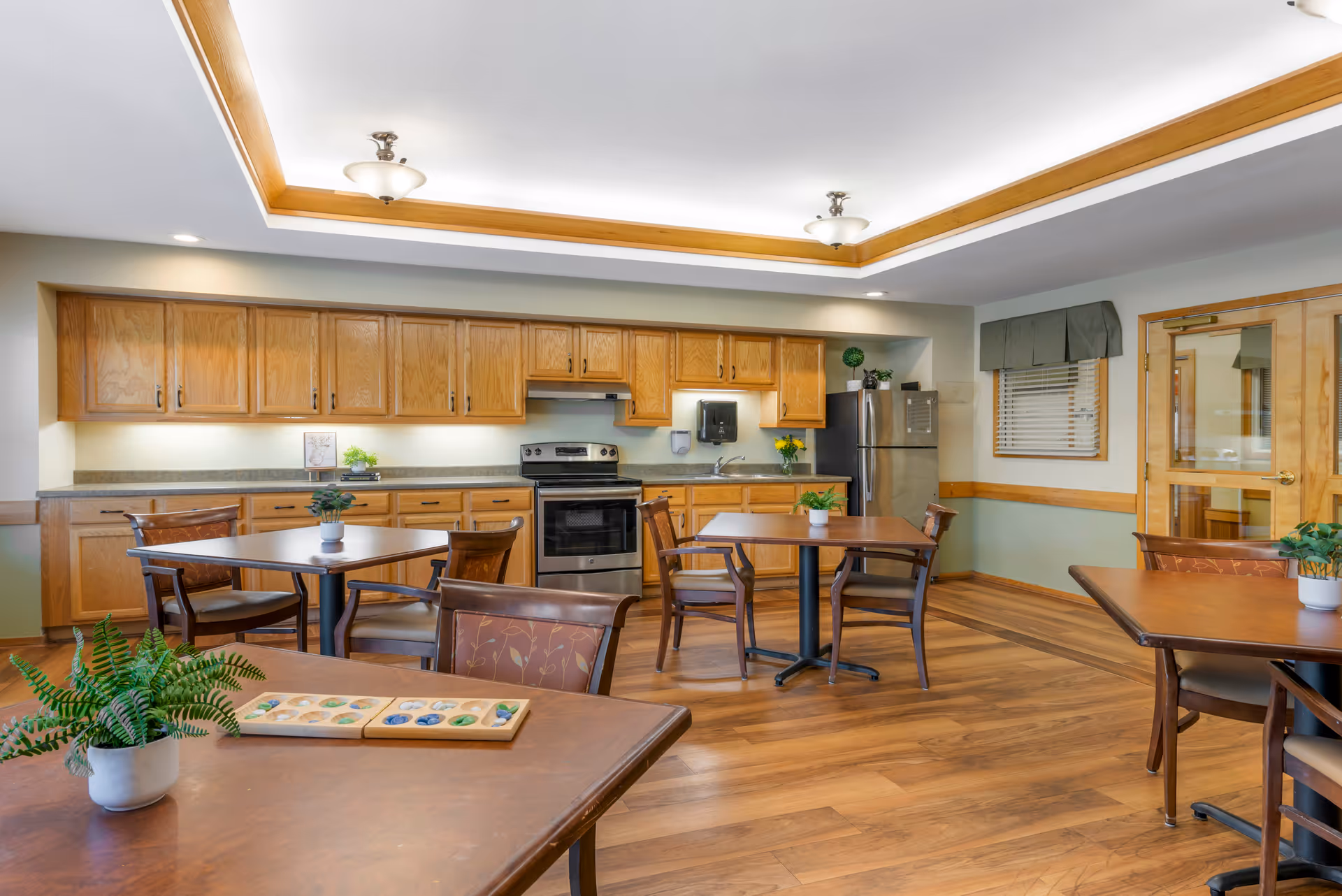 Communal dining area with wooden tables and chairs facing a kitchen with oak cabinets, stove, and refrigerator.