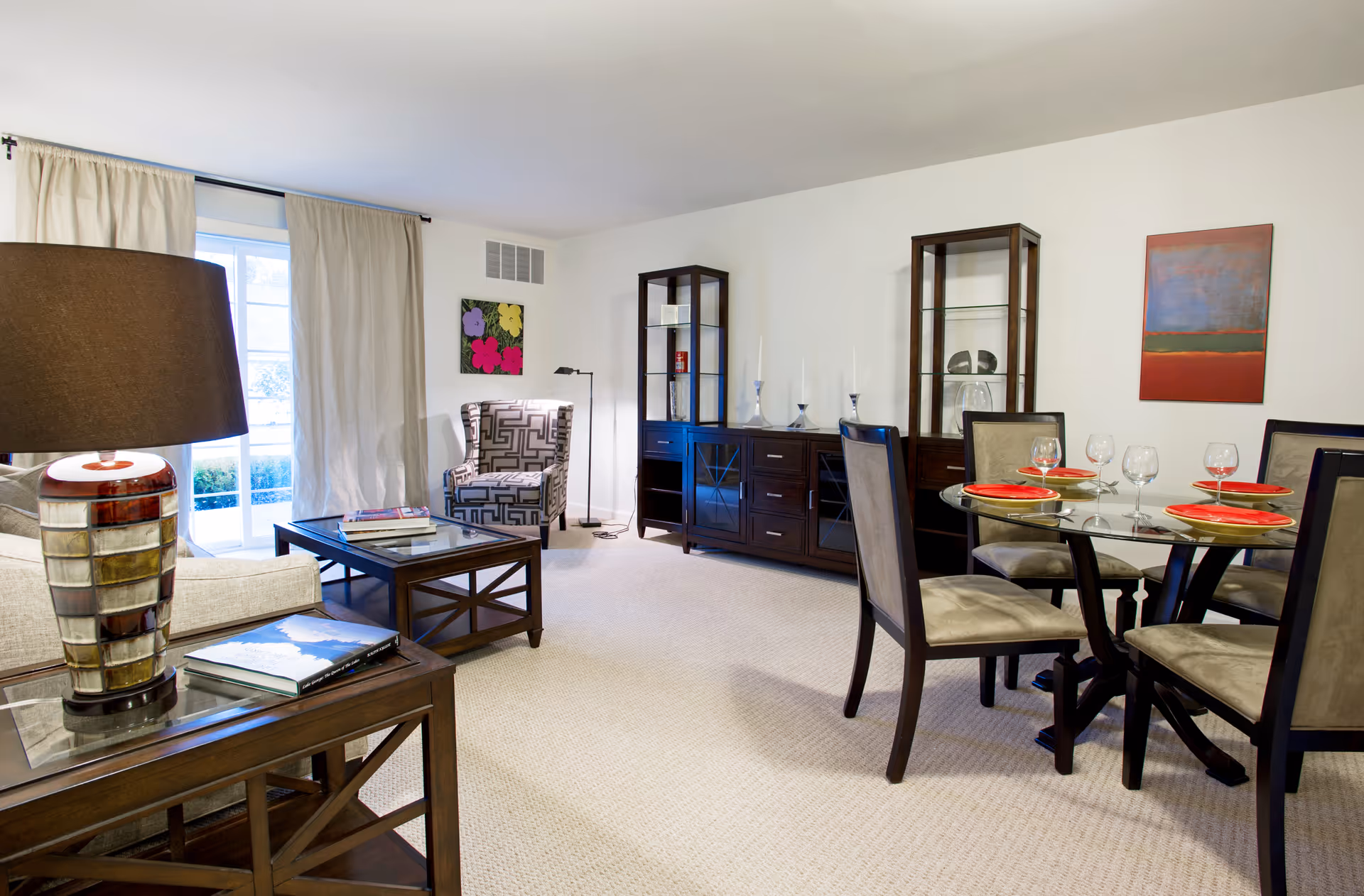 A bright and spacious living and dining area with beige carpet flooring. The room features a glass-top dining table set with four chairs, each with beige upholstery. On the table are red plates and wine glasses. Against the wall are two dark wood display cabinets and a matching sideboard with decorative items on top. A patterned armchair is positioned near a window with beige curtains, and a wooden coffee table and side table with a lamp and books complete the seating area.