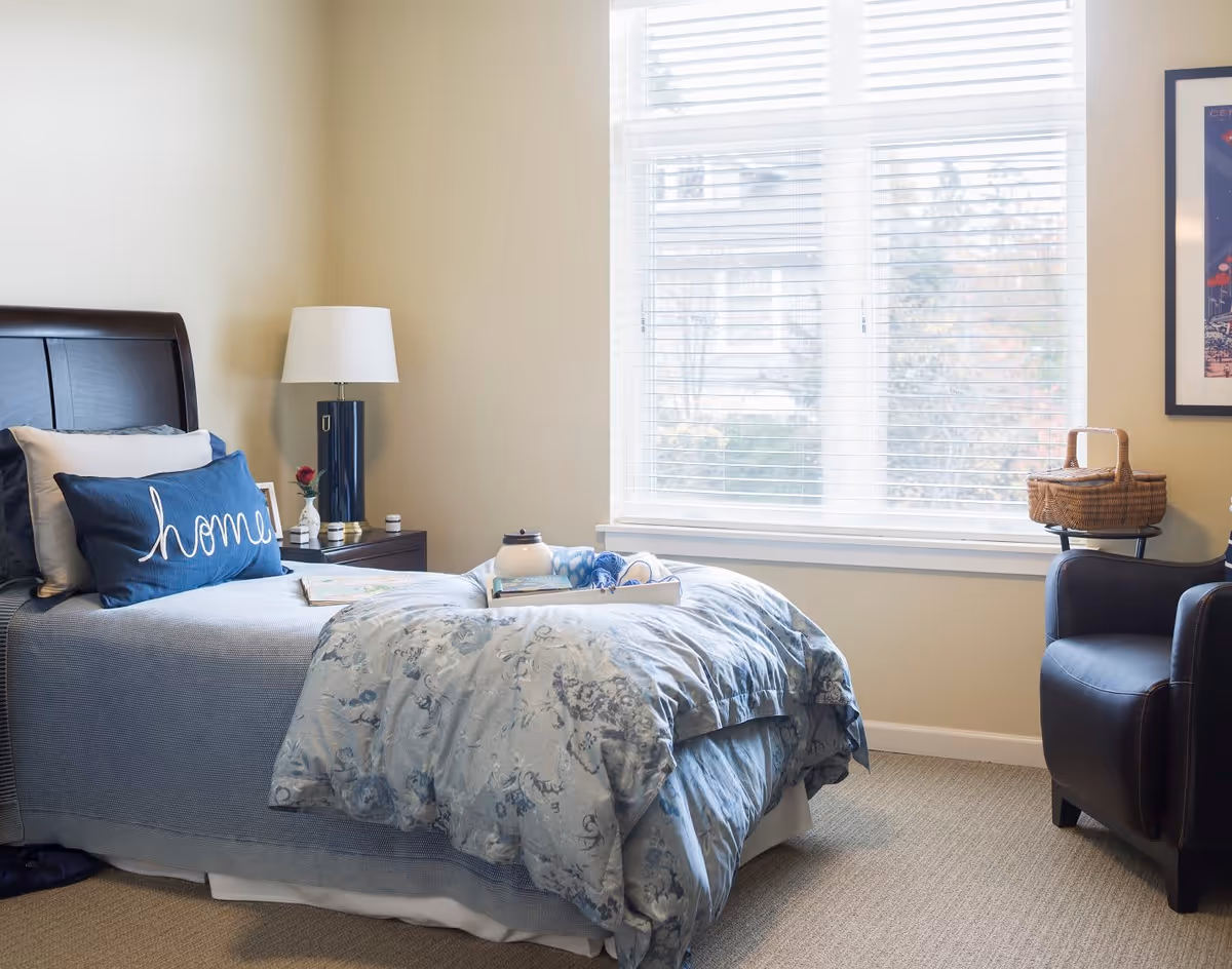 A cozy bedroom with a single bed featuring blue and white bedding, a decorative pillow with the word 'home', a bedside table with a lamp and small decor items, a large window with white blinds letting in natural light, a dark armchair with a wicker basket on it, and a framed picture on the wall.