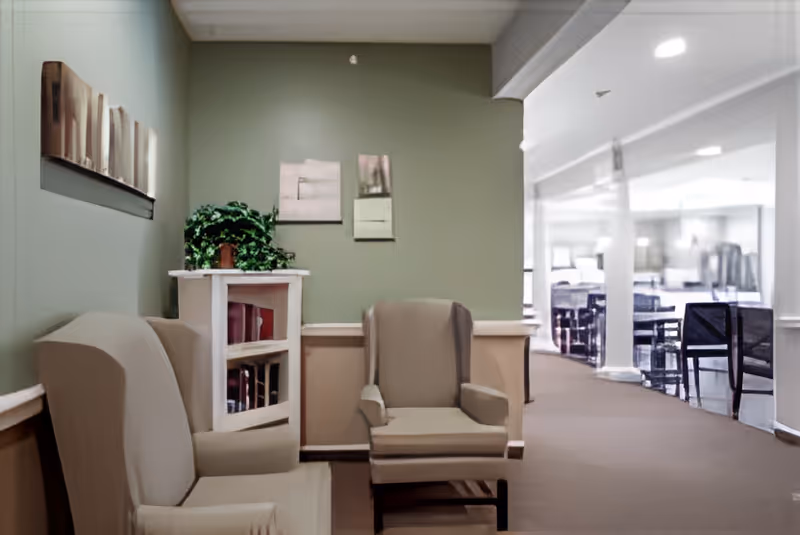 A quiet seating area with two beige armchairs and a small white bookshelf with a green plant on top, set against a green wall with two small framed pictures. In the background, there is a hallway leading to a dining area with tables and chairs.