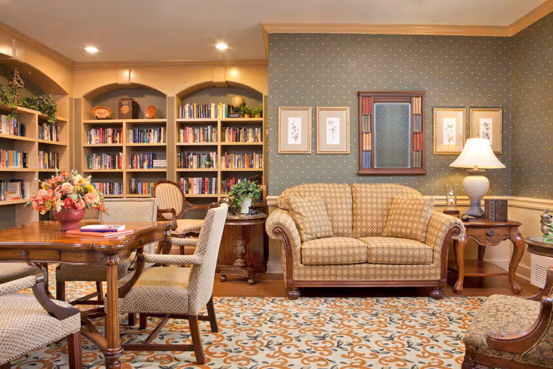 A cozy living room area with a patterned sofa, wooden side tables with a lamp, and framed artwork on the wall. There is a wooden table with upholstered chairs and a floral centerpiece. In the background, built-in bookshelves filled with books and decorative items are visible. The room has warm lighting and a patterned carpet.