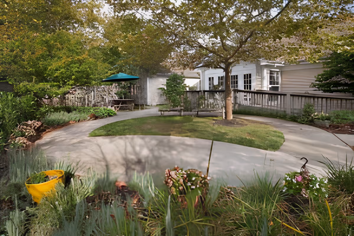 A peaceful outdoor garden area with a curved concrete pathway surrounding a grassy patch with a tree in the center. There are benches along the path, various plants and flowers in the foreground, and a picnic table with a green umbrella in the background near a wooden fence and white building.