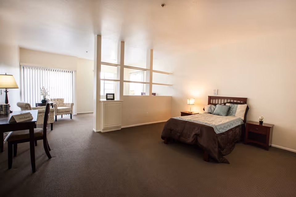 A spacious retirement center room with a bed on the right side, two nightstands with lamps, a small table with chairs on the left, and two armchairs near a large window with vertical blinds. The room has beige walls and carpeted floor, with a partial divider featuring open shelves separating the bed area from the sitting area.