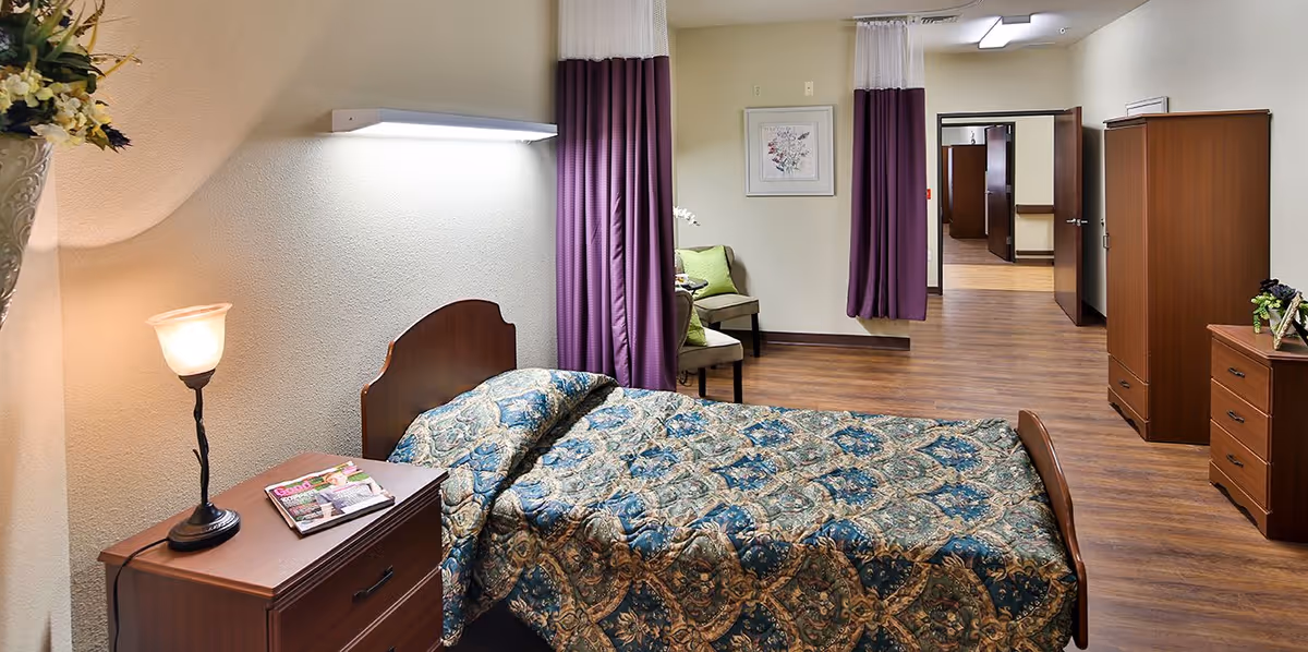 A senior living facility bedroom with a single bed covered in a patterned blue and beige bedspread. Next to the bed is a wooden nightstand with a lamp and magazines on it. The room has wooden flooring, purple curtains, a chair with green cushions, and wooden dressers. The walls are light-colored and there is a framed floral picture hanging on the wall.