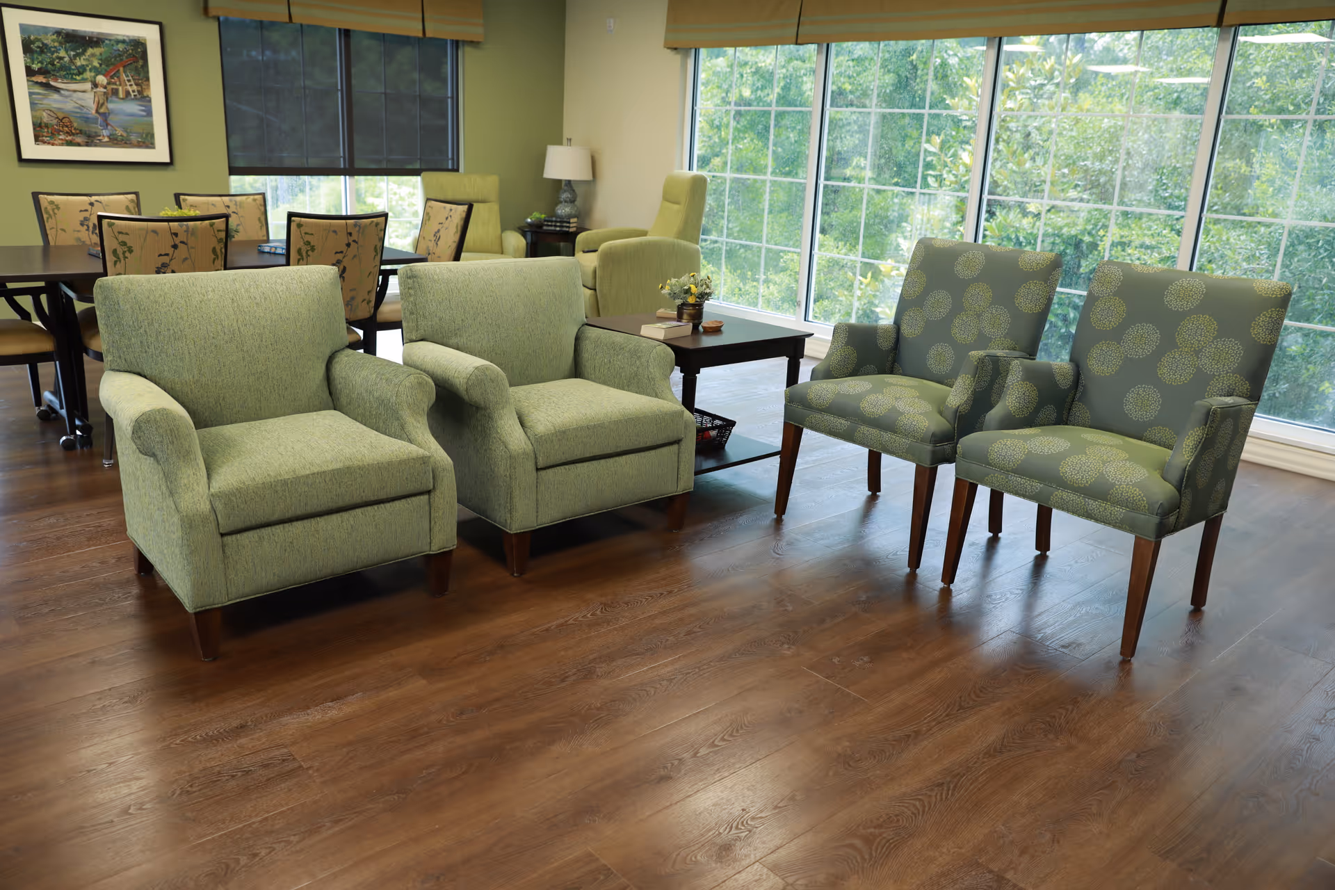 A bright and spacious common area with four green upholstered chairs arranged around a wooden coffee table. Large windows in the background let in natural light and offer a view of greenery outside. In the background, there is a dining table with chairs and a framed picture on the wall.