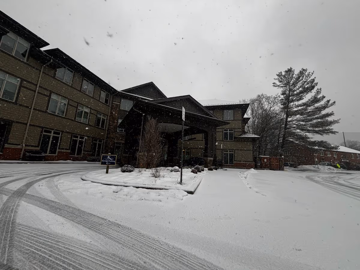Snow-covered driveway and entrance of a multi-story senior living building with a covered porte-cochere and surrounding trees.