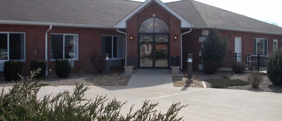 Front exterior view of a single-story brick building with a central entrance featuring double glass doors and an arched window above. The entrance is flanked by small bushes and outdoor lighting fixtures, with a concrete walkway leading up to it.