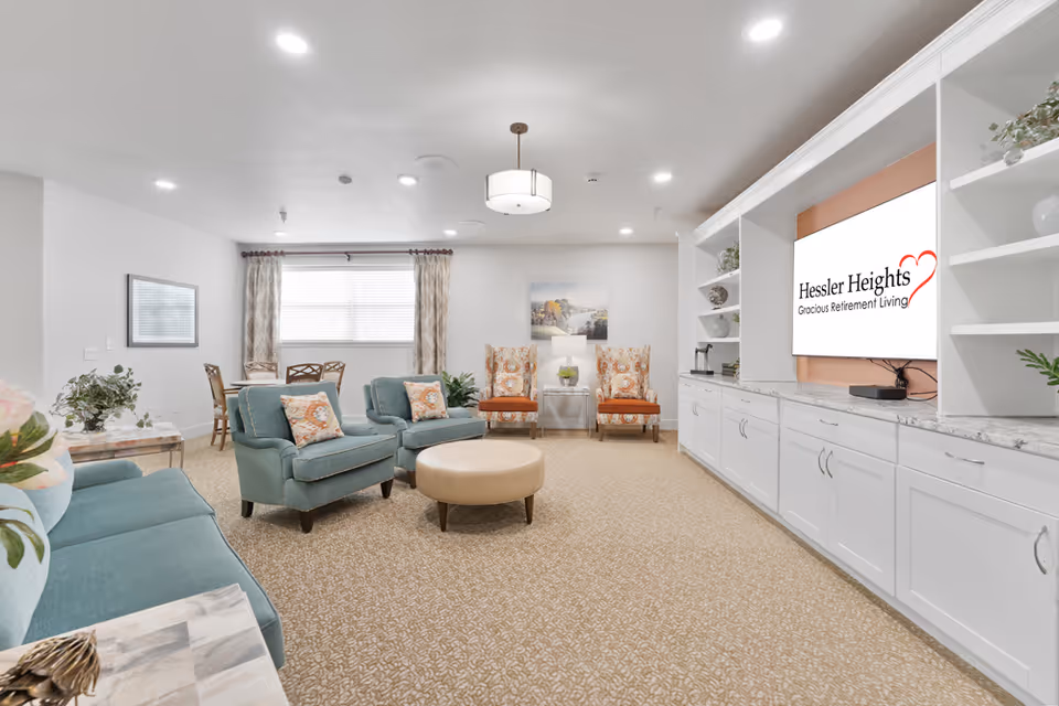A bright and spacious living room in a retirement facility featuring teal upholstered sofas and armchairs with patterned cushions, two orange patterned armchairs, a round beige ottoman, a large built-in white cabinet with shelves and a mounted TV displaying the Hessler Heights Gracious Retirement Living logo, beige patterned carpet, and a window with curtains.