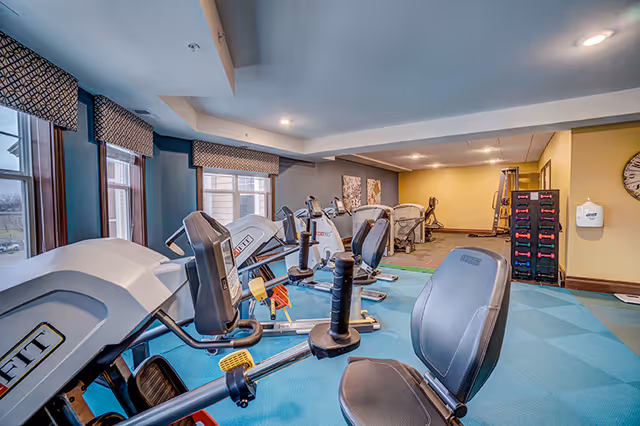 Interior view of a fitness room in a senior living facility with several exercise machines including recumbent bikes and ellipticals. The room has blue carpet flooring, large windows with patterned valances, and a rack of colorful dumbbells against a yellow wall.