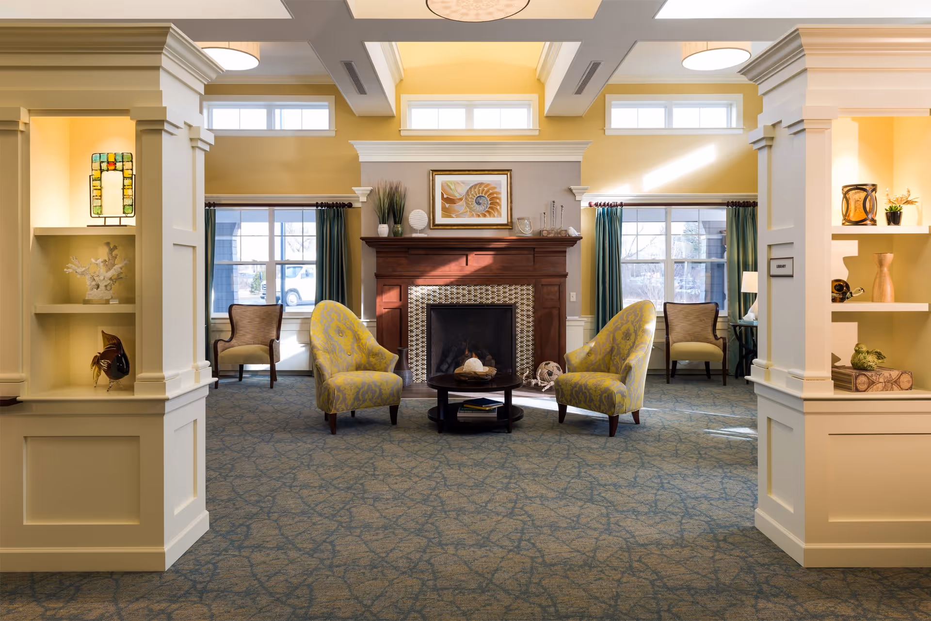 A cozy living room area with two yellow patterned armchairs facing a dark wooden coffee table in front of a fireplace with a decorative tile surround. The room has large windows with teal curtains, built-in shelves with decorative items, and soft yellow walls with white trim and columns. Natural light fills the space, creating a warm and inviting atmosphere.