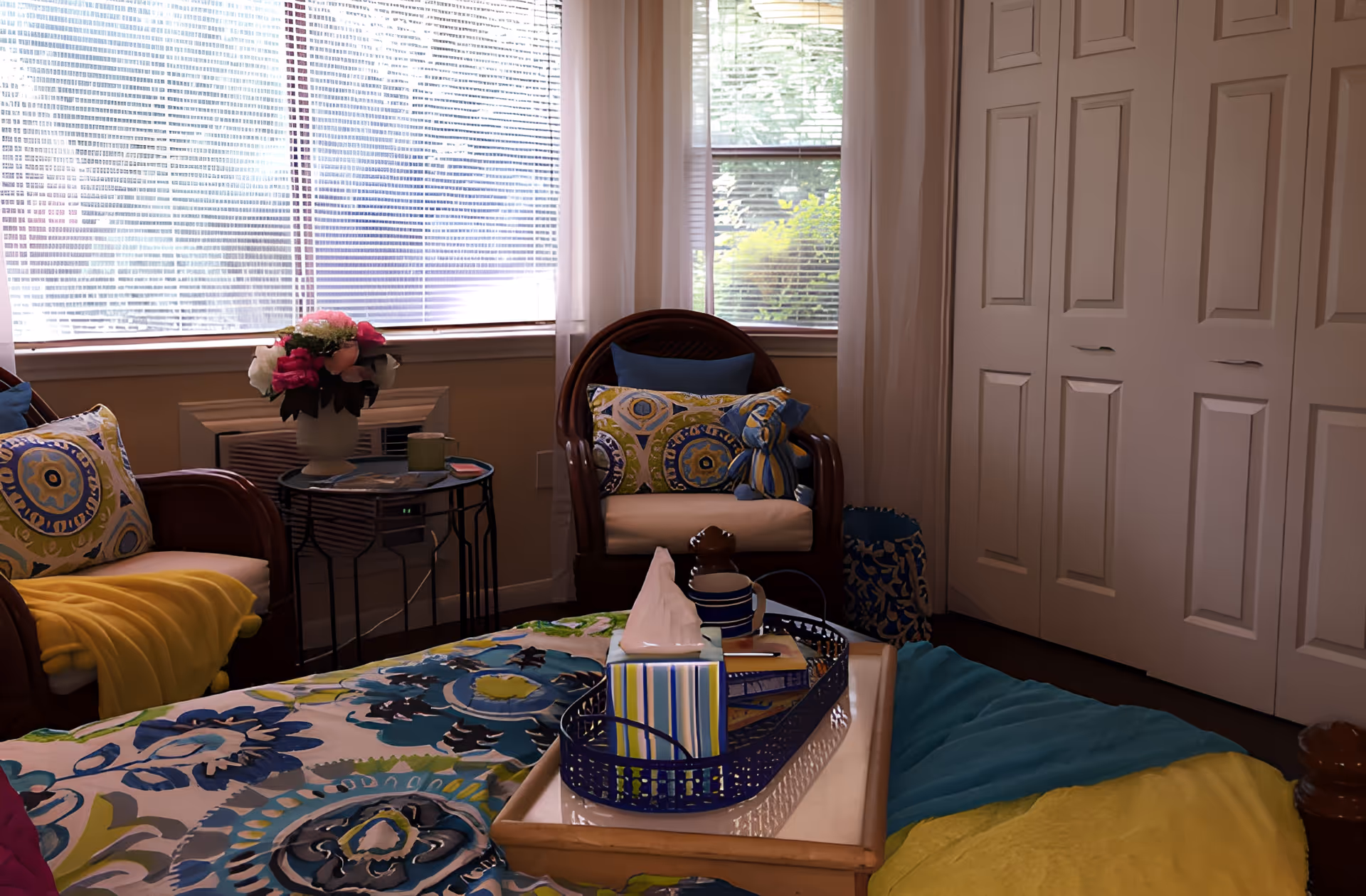 Cozy bedroom with a bed covered in blue-green floral bedding, a tray with tissues on it, wicker chairs with patterned pillows, a side table with flowers, and a window with blinds.