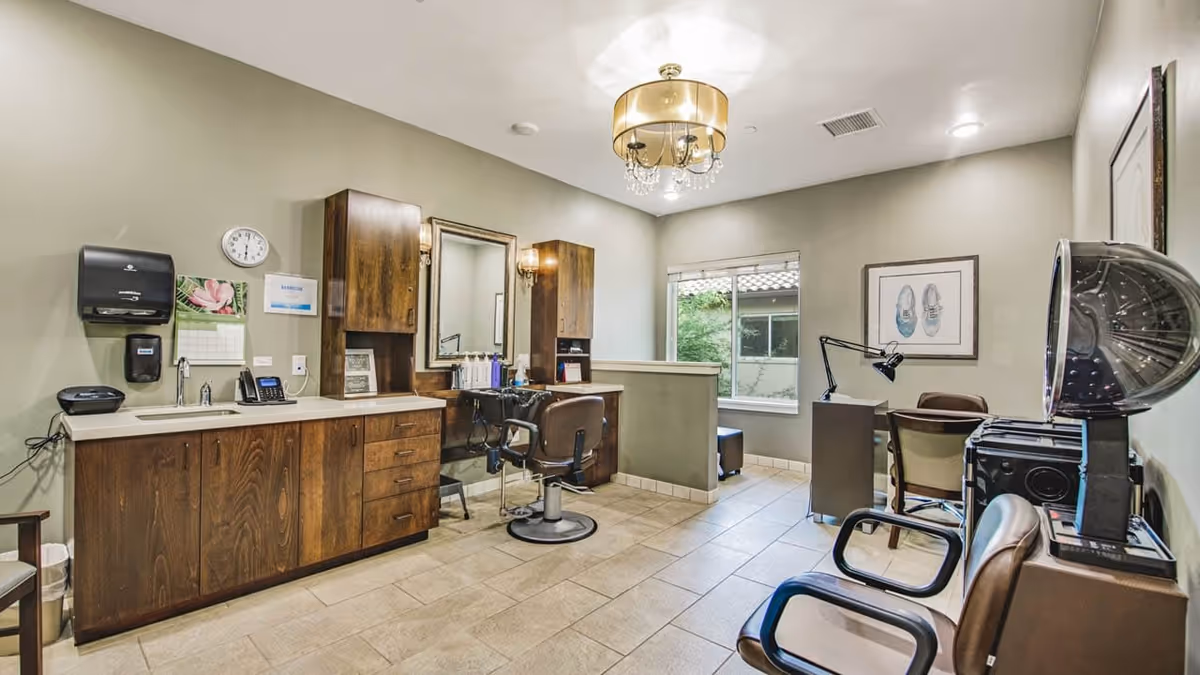 Interior view of a salon area in a senior living facility featuring a styling chair in front of a mirror with wooden cabinets, a sink, and hair care products. There is a hair dryer chair on the right and a desk with a lamp and chair near a window showing greenery outside. The room has beige tiled flooring and soft lighting from a ceiling chandelier.