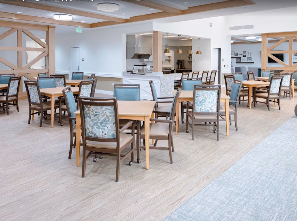 A bright and spacious dining area with multiple wooden tables and chairs featuring floral and solid blue upholstery. The room has light wood flooring, a ceiling with wooden beams, and modern light fixtures. In the background, there is a kitchen area with a granite countertop and pendant lights.